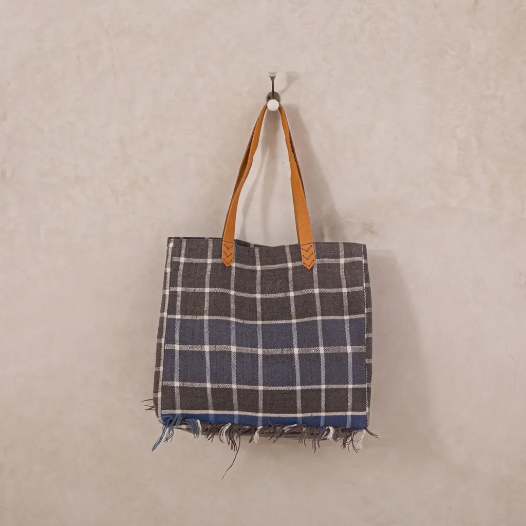 A plaid-patterned tote bag with brown leather handles hangs on a wall hook against a beige textured backdrop, perfect for adding charm to any room.