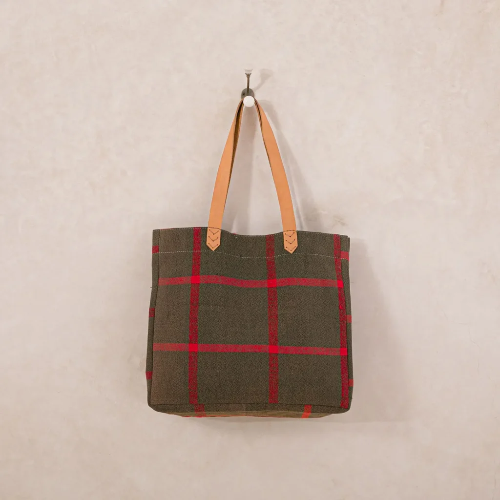 Tote bag with red and green plaid design, tan leather handles, hanging on a hook against a beige wall.