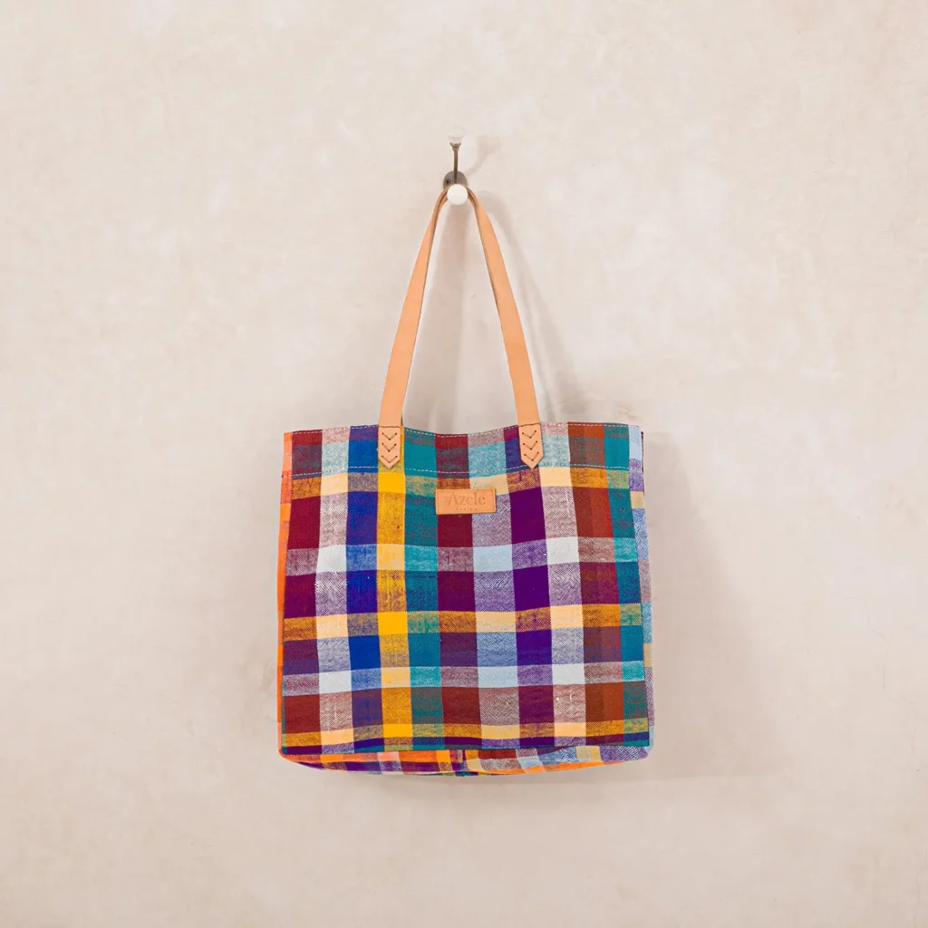 A colorful plaid tote bag with leather handles hangs on a wall hook.