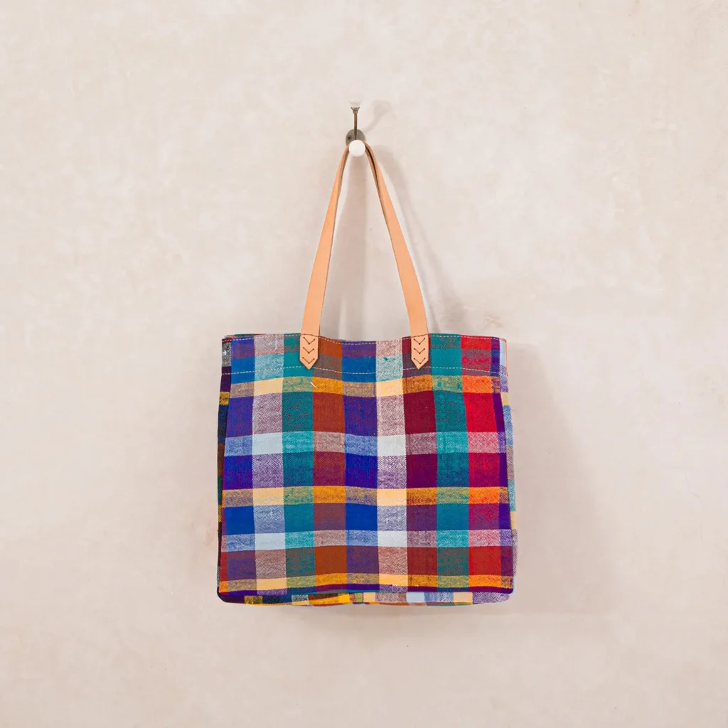 A colorful plaid tote bag with tan handles hangs on a wall hook.