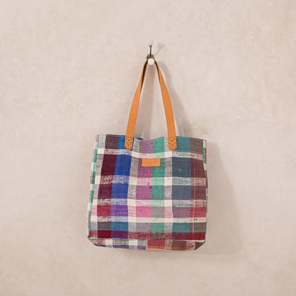 A multicolored plaid tote bag with brown leather handles is hanging on a wall hook.