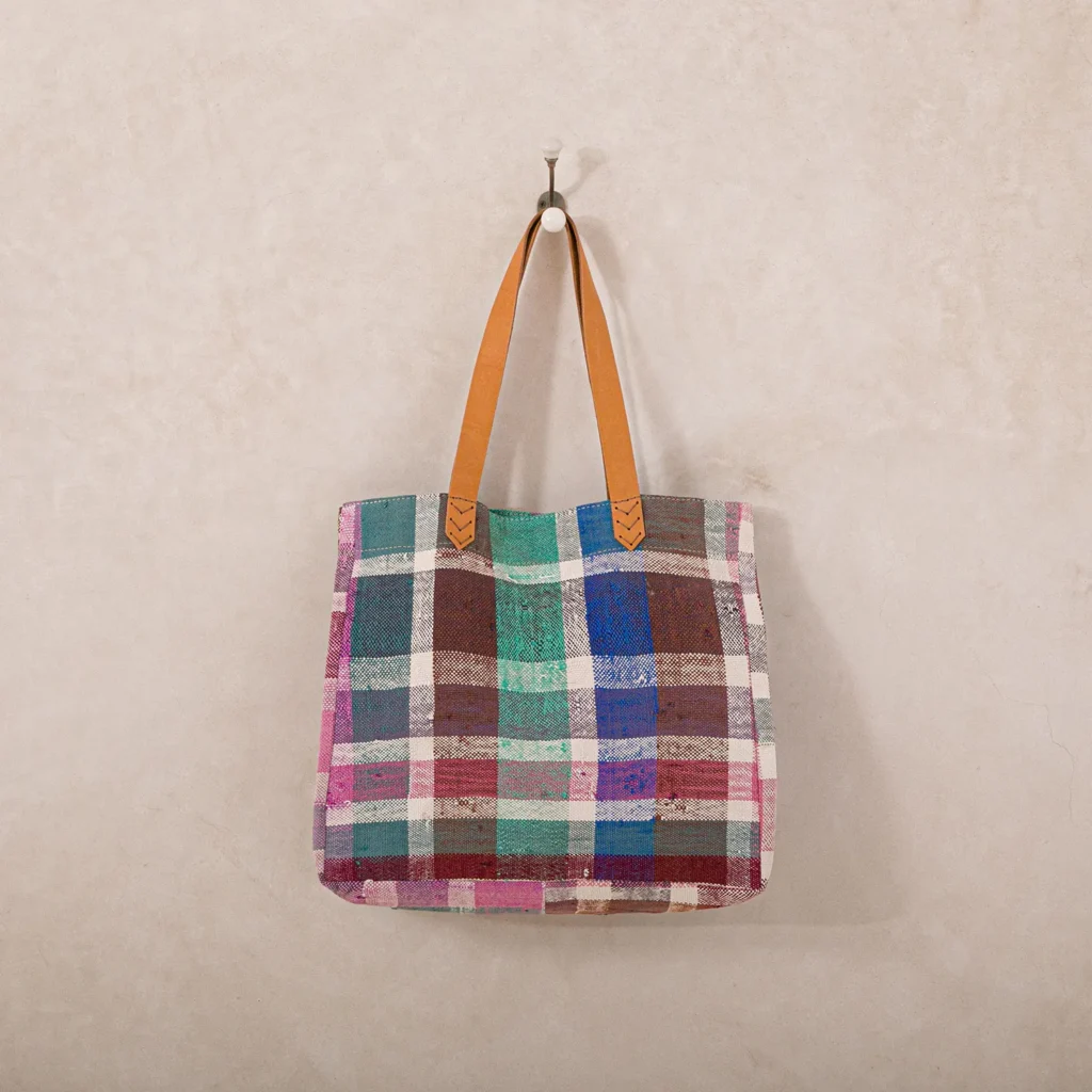 A multicolored patchwork tote bag with tan leather handles hanging on a peg against a plain wall.