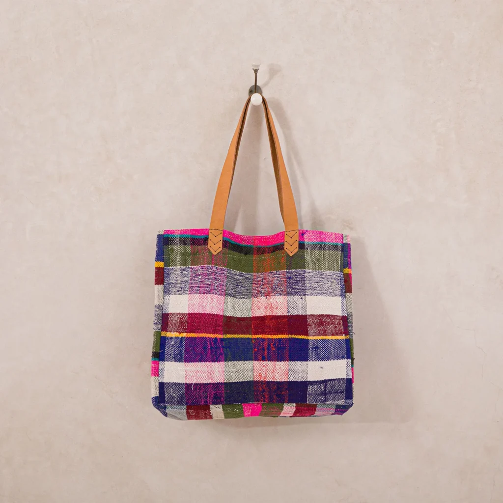 Colorful plaid tote bag with brown leather handles hanging on a white wall hook.