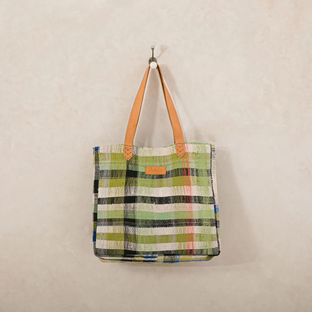 A plaid tote bag with green, black, and white stripes hangs on a wall hook. It features tan leather handles and a brown label on the front.