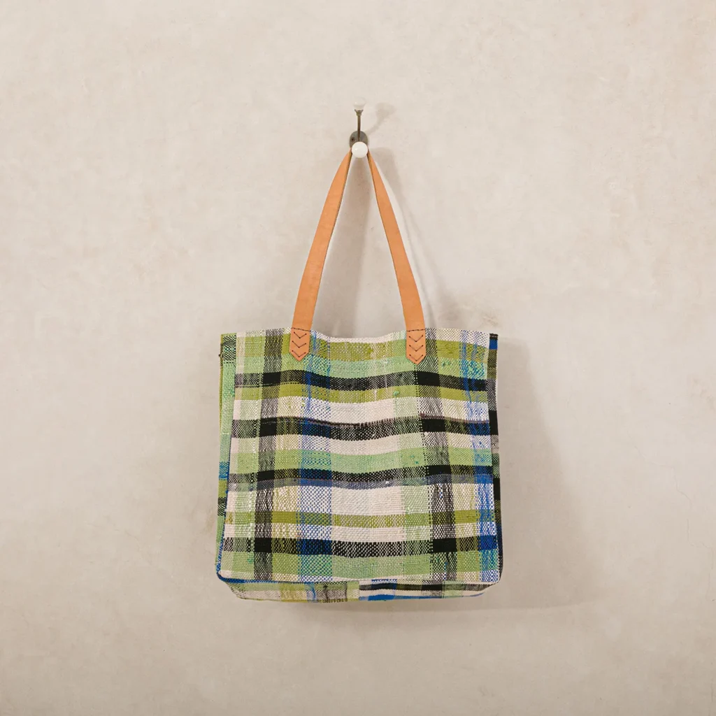 Green and blue checkered tote bag with tan leather straps hanging on a wall hook.