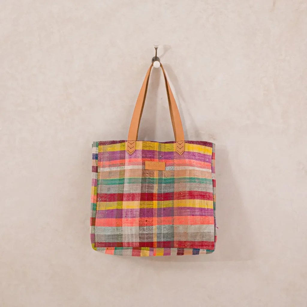 A multicolored plaid tote bag with brown leather handles hangs on a wall hook against a beige background.