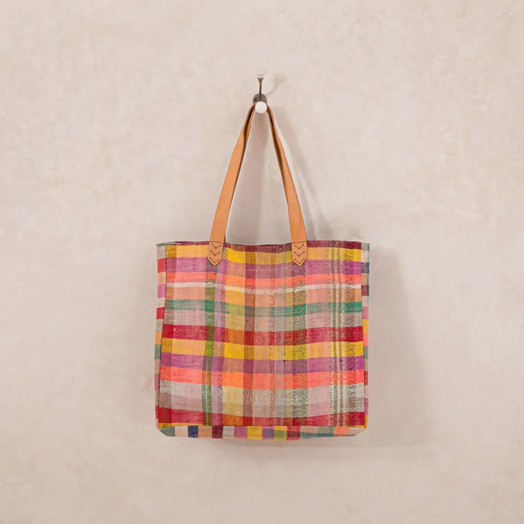 A plaid multicolored tote bag with tan leather straps hangs on a wall hook.