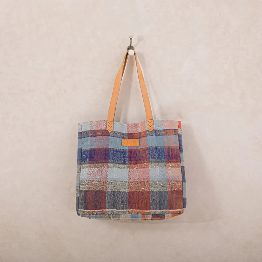 A plaid-patterned tote bag with brown leather handles hangs on a wall hook against a plain beige background.
