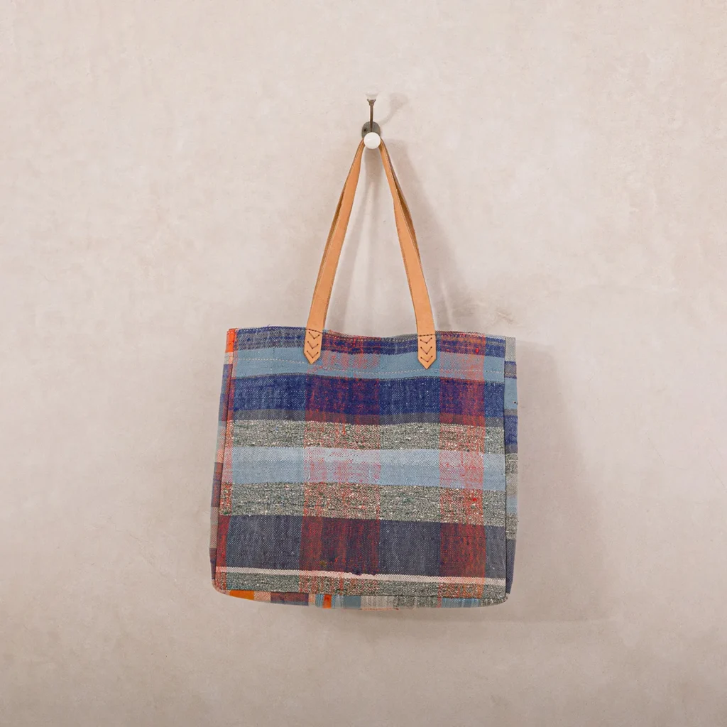 A multicolored striped tote bag with brown leather handles hangs on a wall hook.