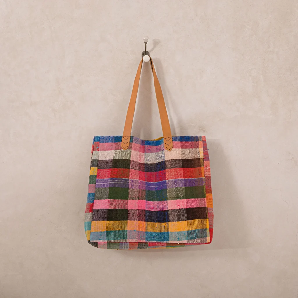 A multi-colored checkered tote bag with tan handles hangs on a single wall hook against a neutral background.
