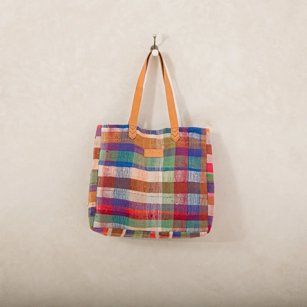 A multicolored plaid tote bag with tan leather handles hangs on a wall hook against a plain backdrop.