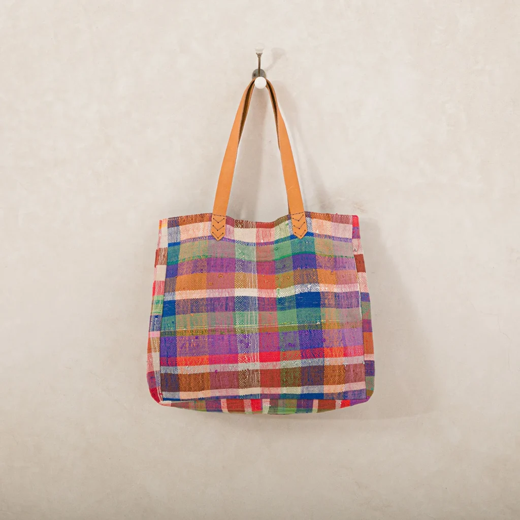 A colorful plaid tote bag with leather straps hangs on a wall hook.