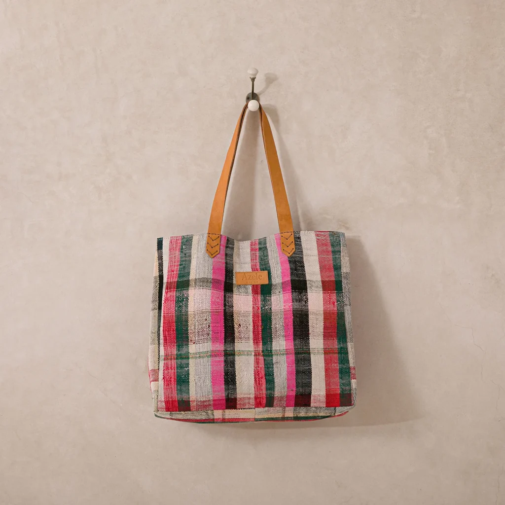 A multicolored plaid patterned tote bag with tan leather handles hangs on a wall hook.