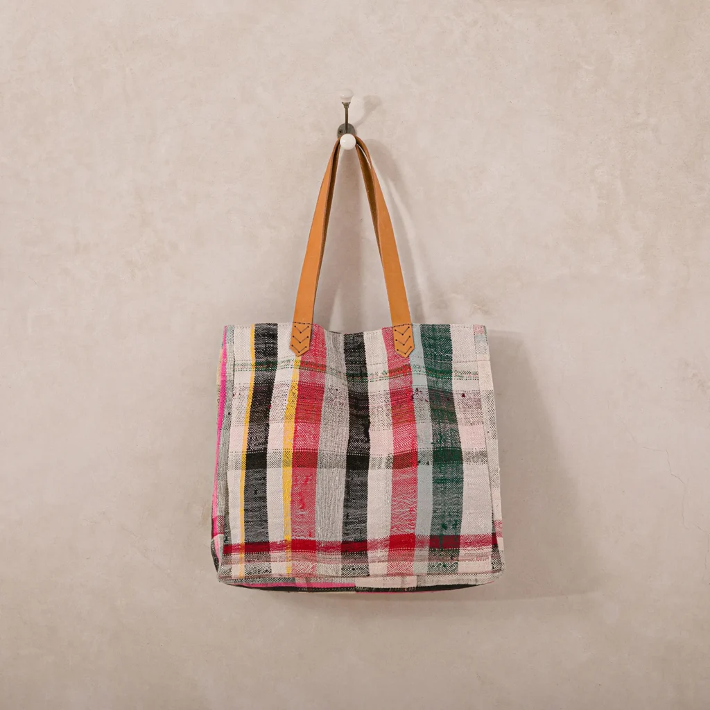 Colorful plaid fabric tote bag with brown leather straps hanging on a wall hook.