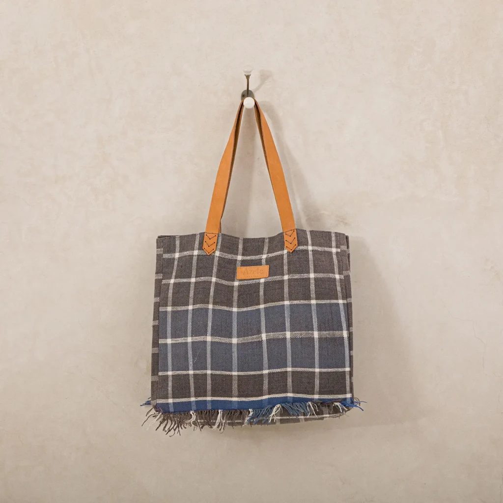 A plaid tote bag with brown leather handles hangs on a wall hook. The bag features a checked pattern and frayed edges at the bottom.