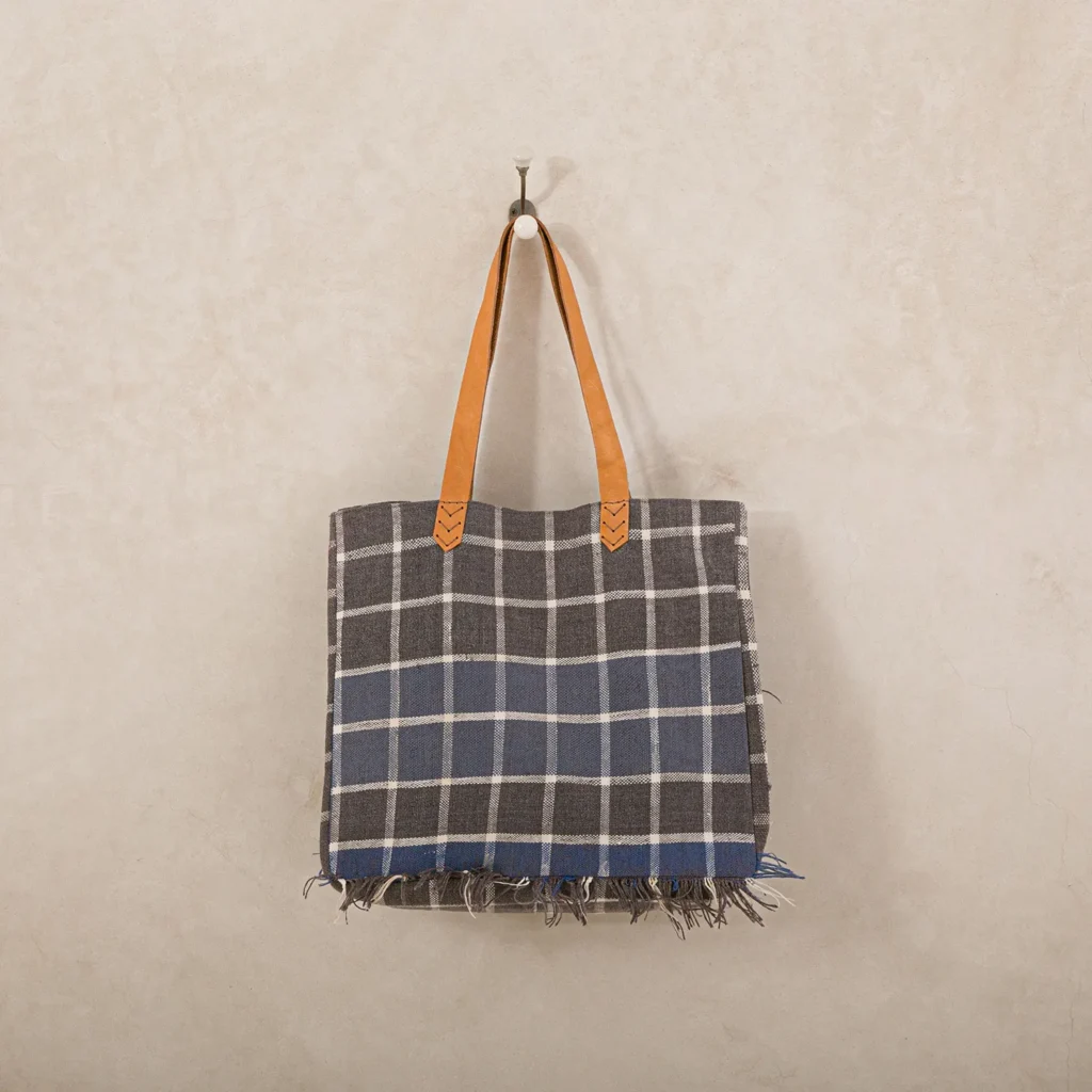 A plaid tote bag with brown leather handles hangs on a wall hook.