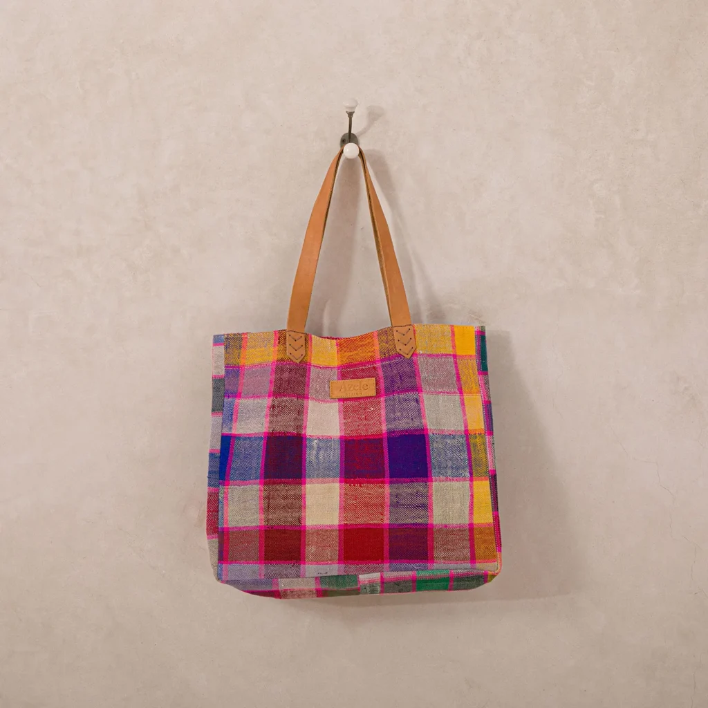 Colorful checkered tote bag with leather straps hanging on a hook against a plain wall.