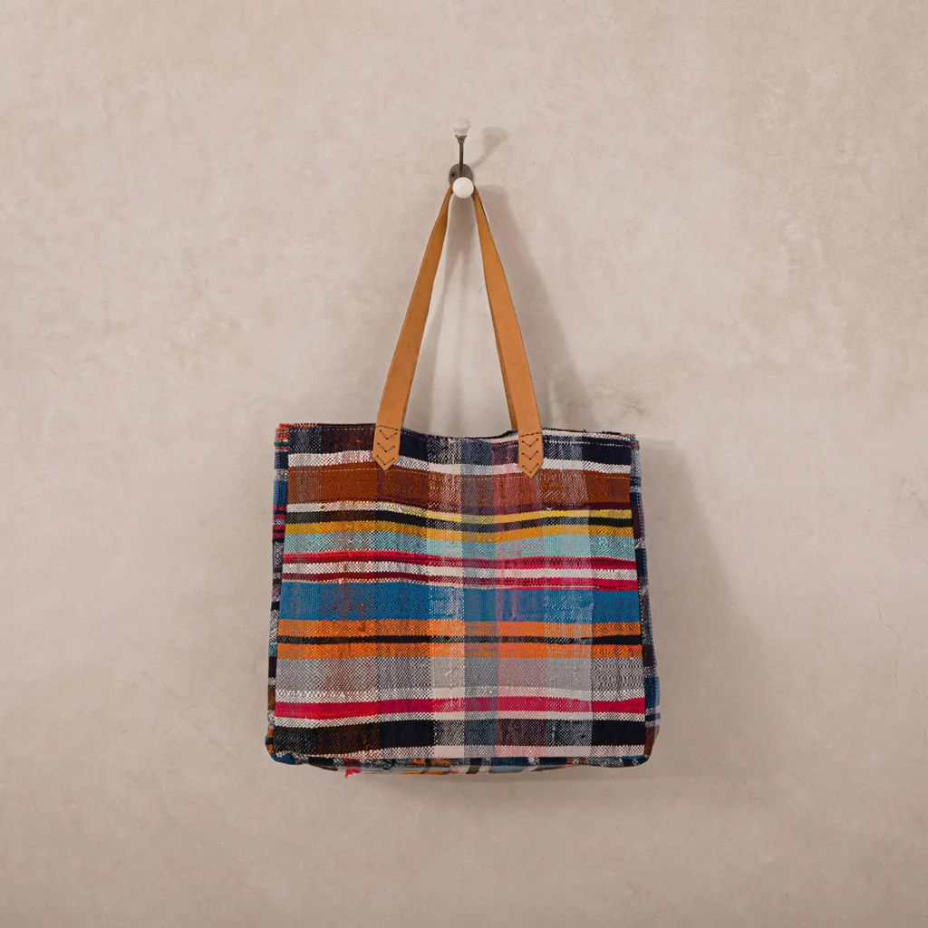 Multicolored striped tote bag with leather handles hanging on a single white wall hook against a beige wall.