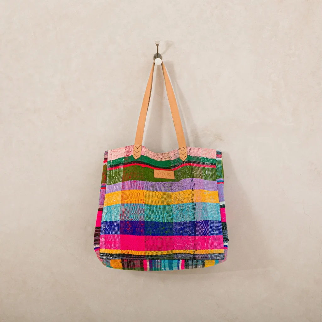 A multicolored woven tote bag with leather handles hangs on a wall hook against a light beige background.