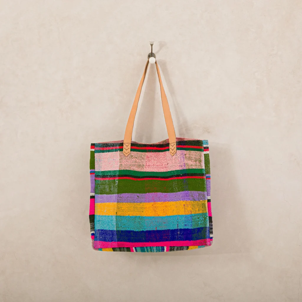 Colorful striped tote bag with tan leather handles hangs on a wall hook.
