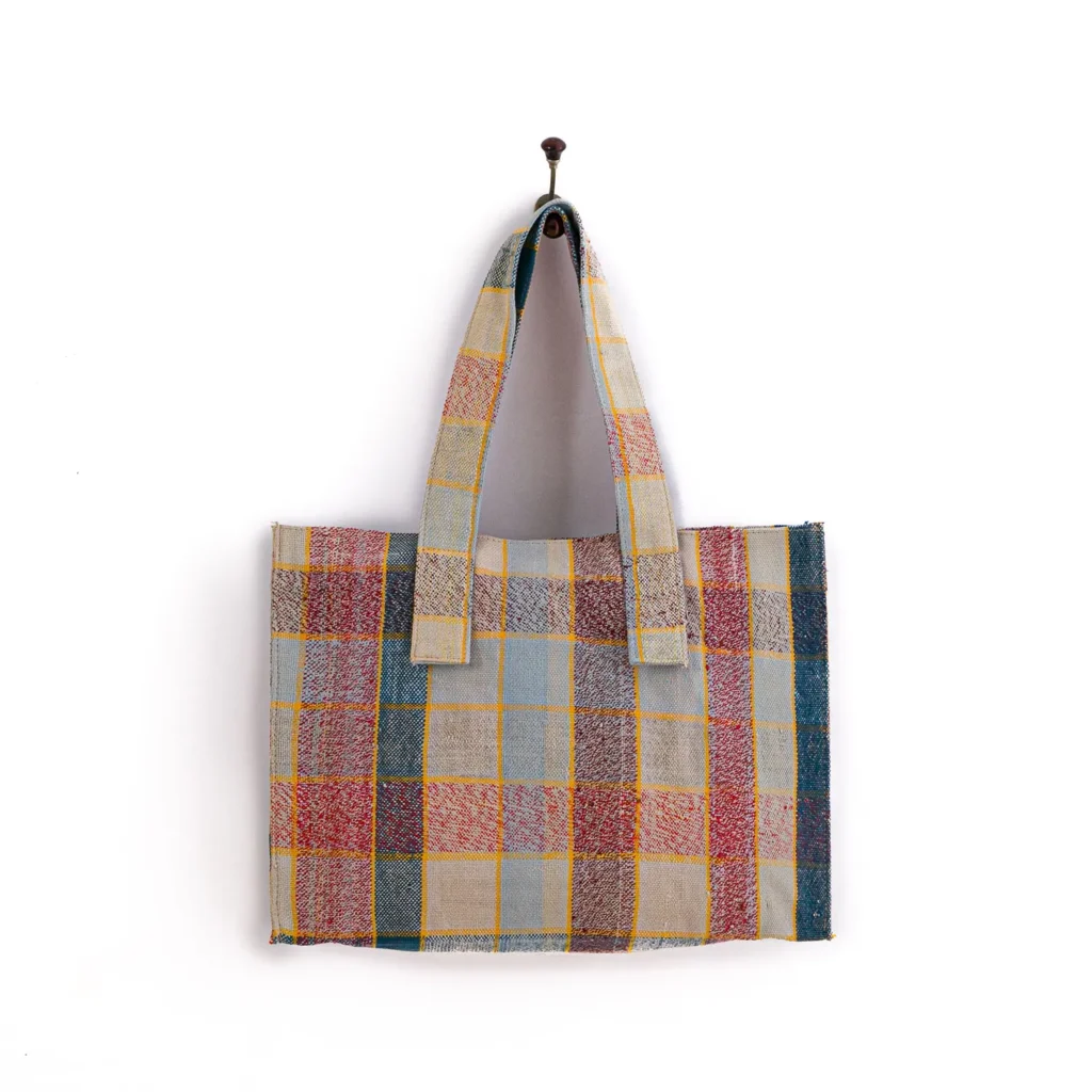 A multicolored plaid tote bag hangs on a wall hook, featuring a checkered pattern in red, yellow, blue, and beige tones.