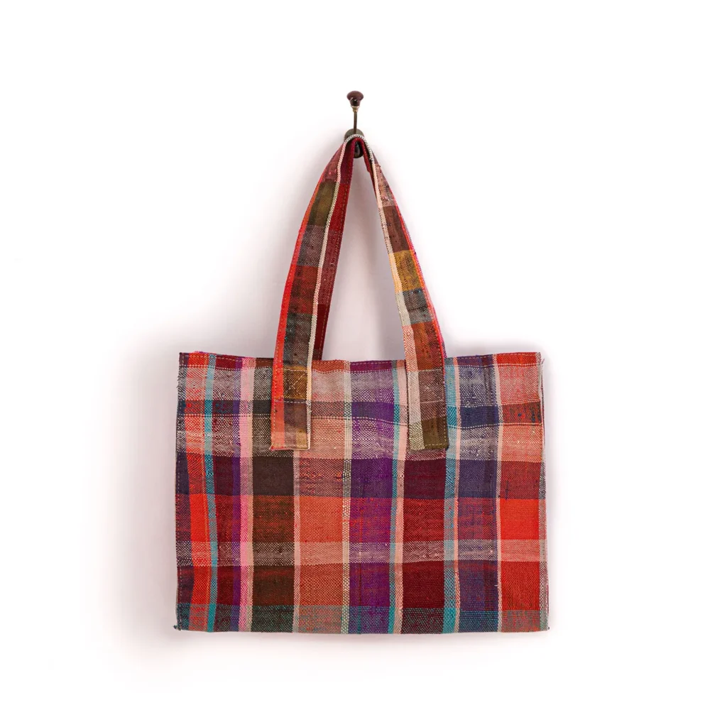 A colorful, plaid-patterned tote bag hangs on a wall hook. The bag features a variety of vibrant, overlapping stripes in red, pink, blue, and orange hues.