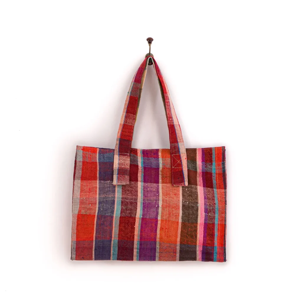 Handmade patchwork bag with a colorful checkered pattern, featuring red, pink, orange, and brown squares, hung on a wall hook.