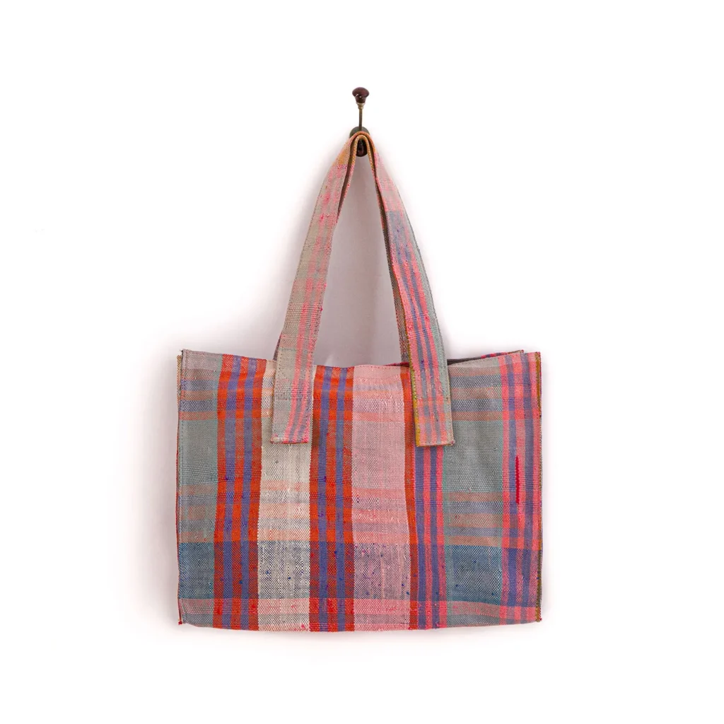 A plaid tote bag with shades of pink, orange, and gray hangs on a wall hook.