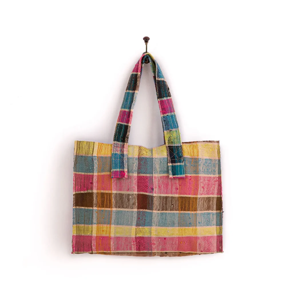Colorful woven tote bag with a checkered pattern of pink, blue, yellow, and brown patches. The bag has two handles and is hanging on a wall hook.