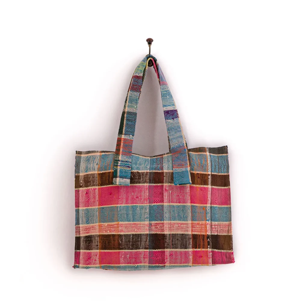 A multicolored, woven tote bag with a checkered pattern hangs on a nail against a plain white wall.