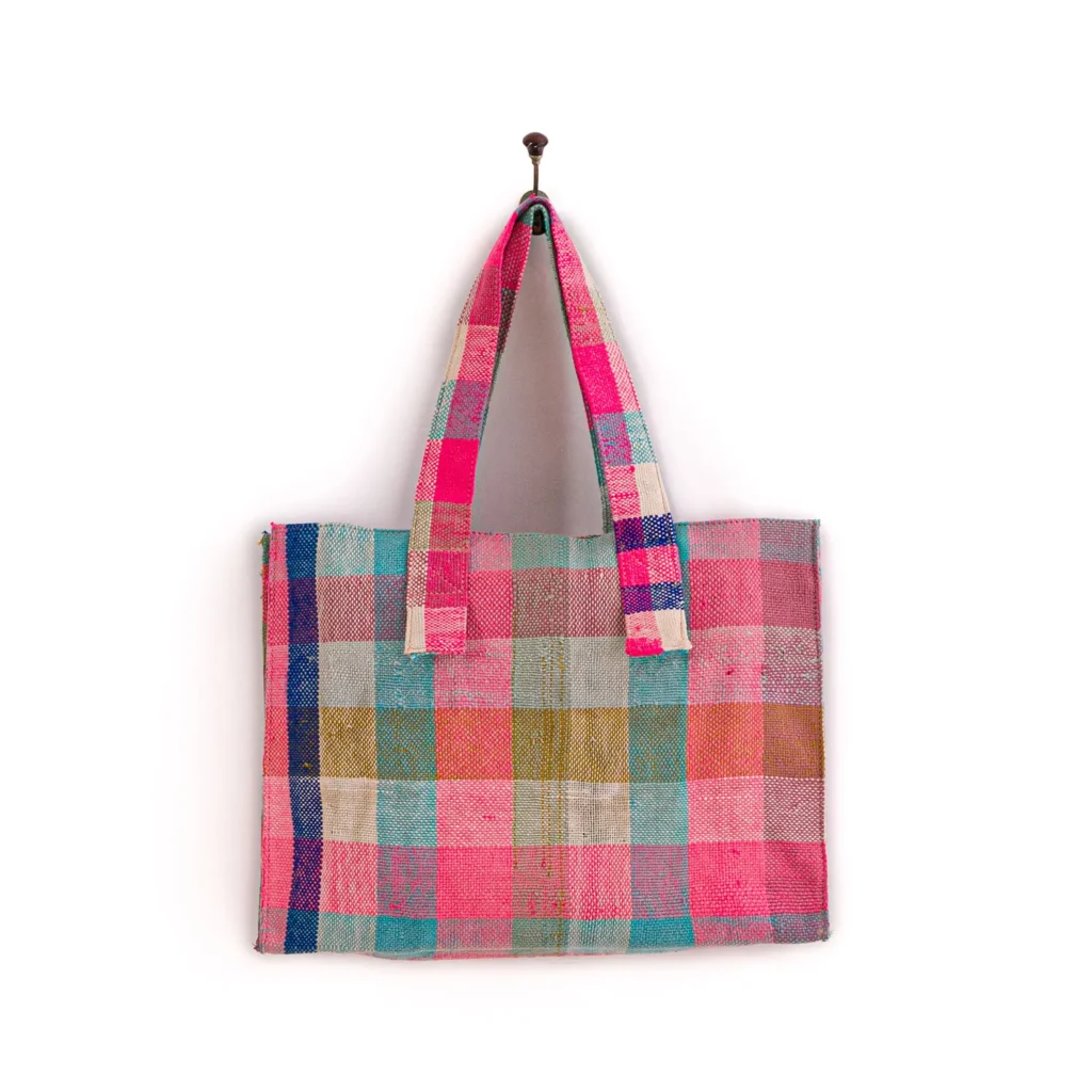 Multicolored, checkered tote bag with pink, blue, and beige patterns hanging on a wall hook.