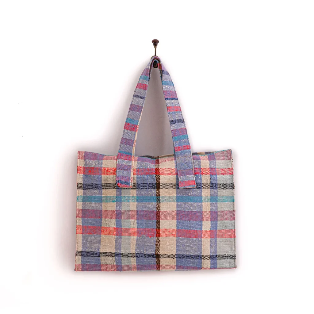 A tote bag with a multicolored plaid pattern, featuring shades of blue, red, and beige, hangs on a hook.