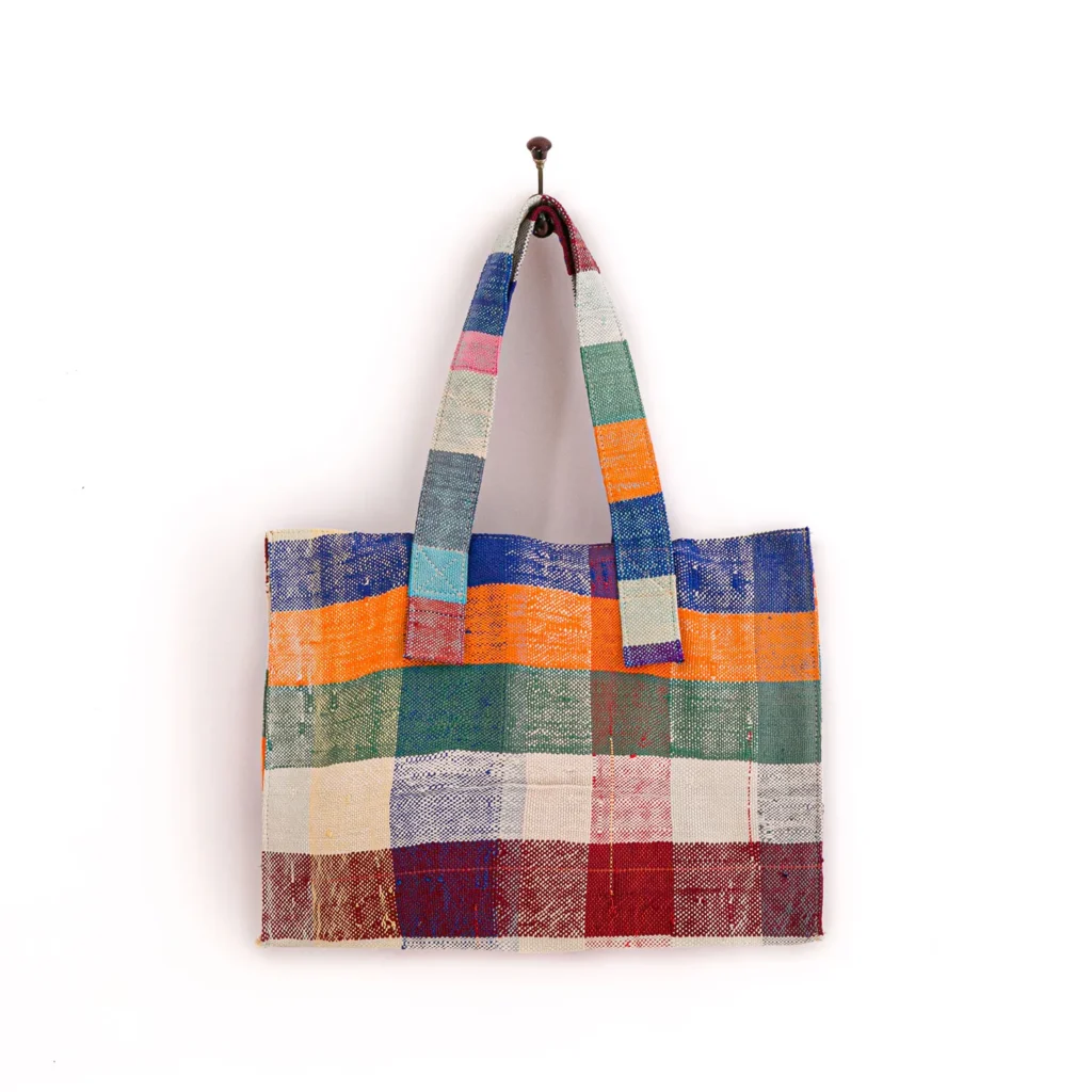A multicolored, checkered tote bag with wide handles hangs on a wall hook against a plain background.