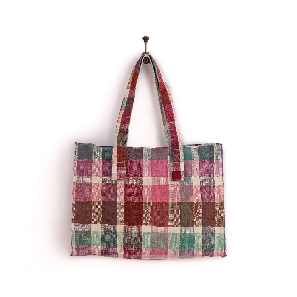 A plaid-patterned tote bag in pink, brown, and green tones, hanging on a wall hook.