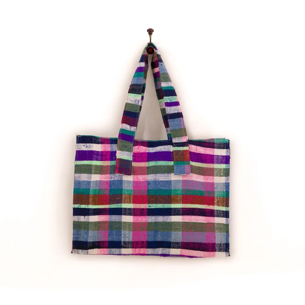 Colorful plaid tote bag with horizontal and vertical stripes in pink, blue, green, and white, hanging on a wall hook.