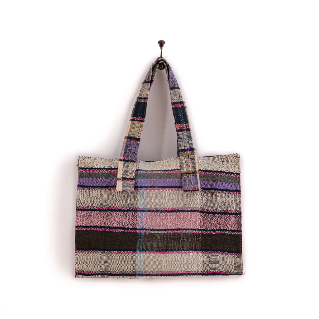 A cloth tote bag with purple, pink, and blue stripes hangs on a single hook against a plain wall.