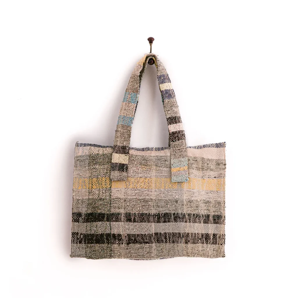 A woven tote bag with a striped pattern in shades of gray, black, and beige, featuring two handles, hanging on a wall hook.