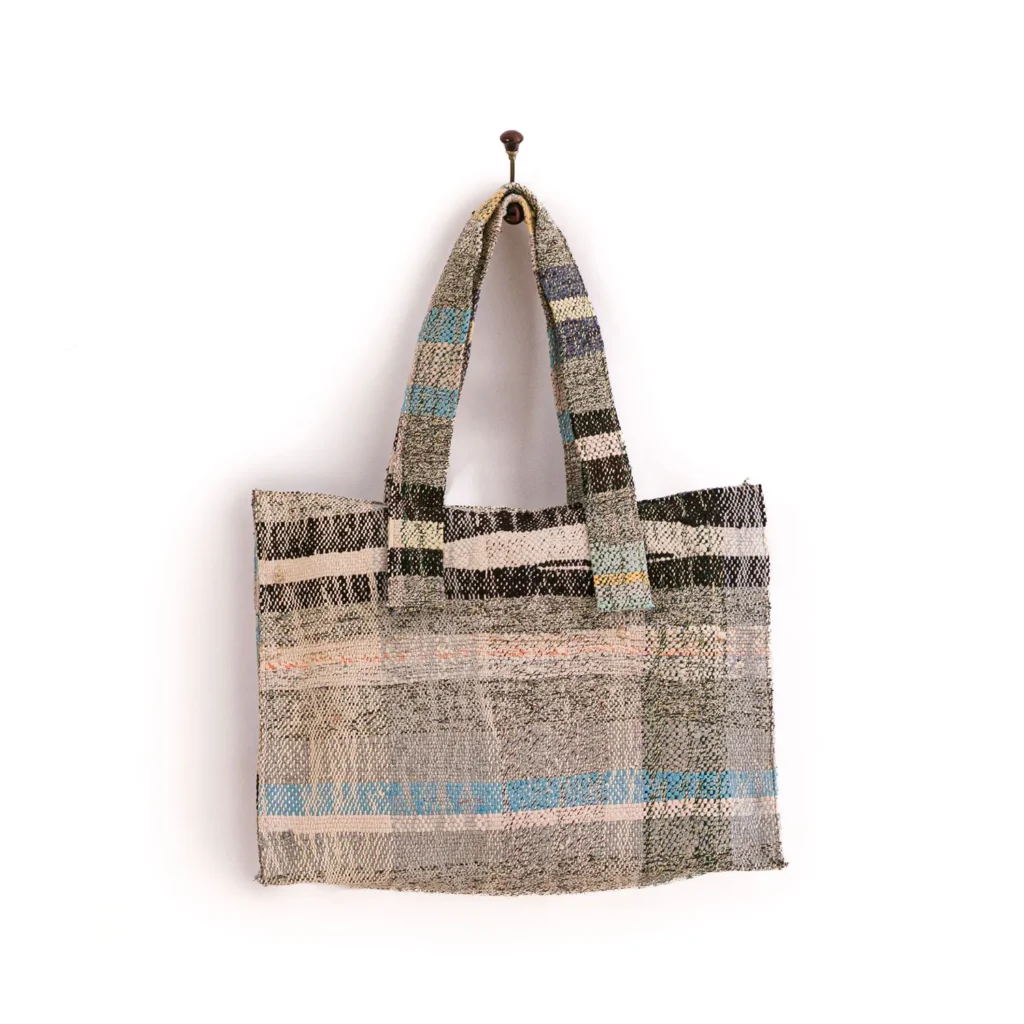 A woven tote bag with a mix of beige, blue, and green tones hangs on a single wall hook.
