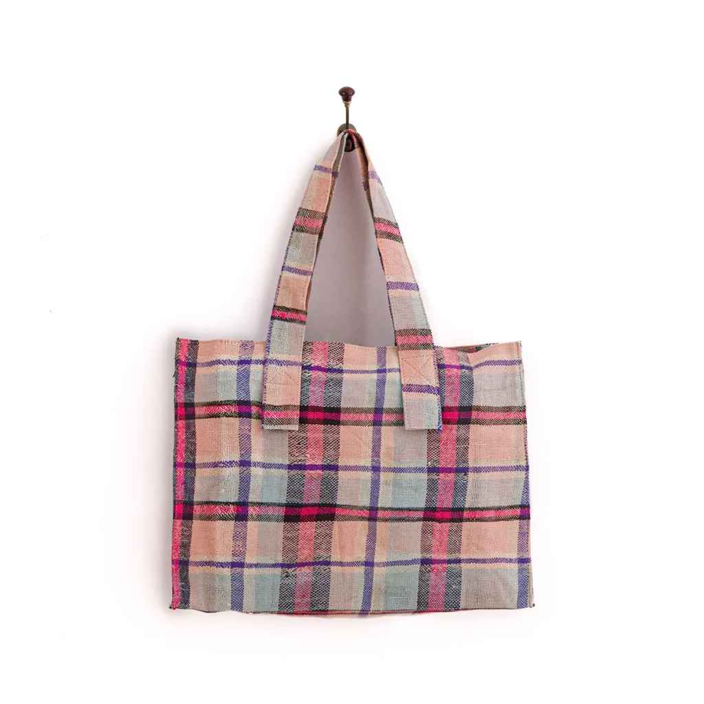A plaid tote bag with pink, blue, and beige stripes hangs from a small hook on a white wall.
