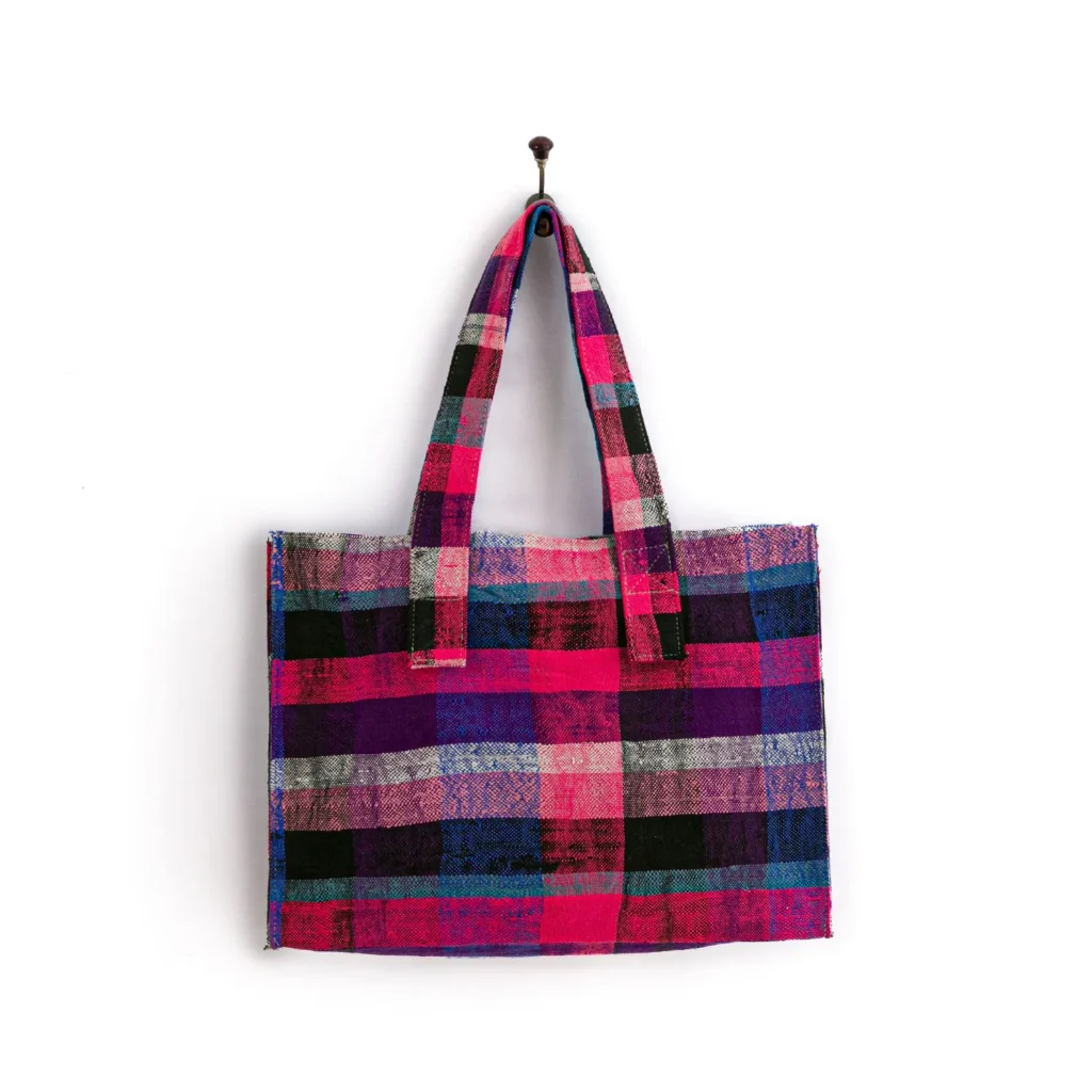 A colorful plaid tote bag with shades of pink, purple, blue, black, and white. The bag has two handles and is hanging on a wall hook.