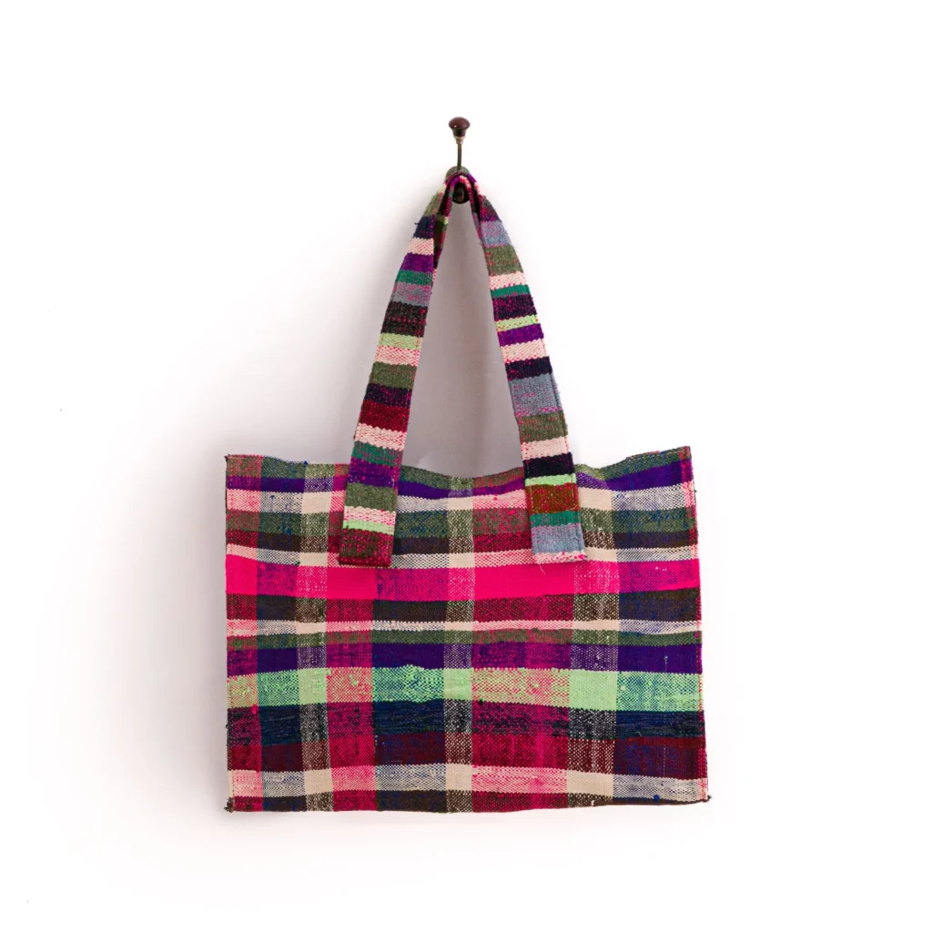 A colorful, checkered woven tote bag with stripes of pink, purple, green, and beige, hanging on a wall hook.