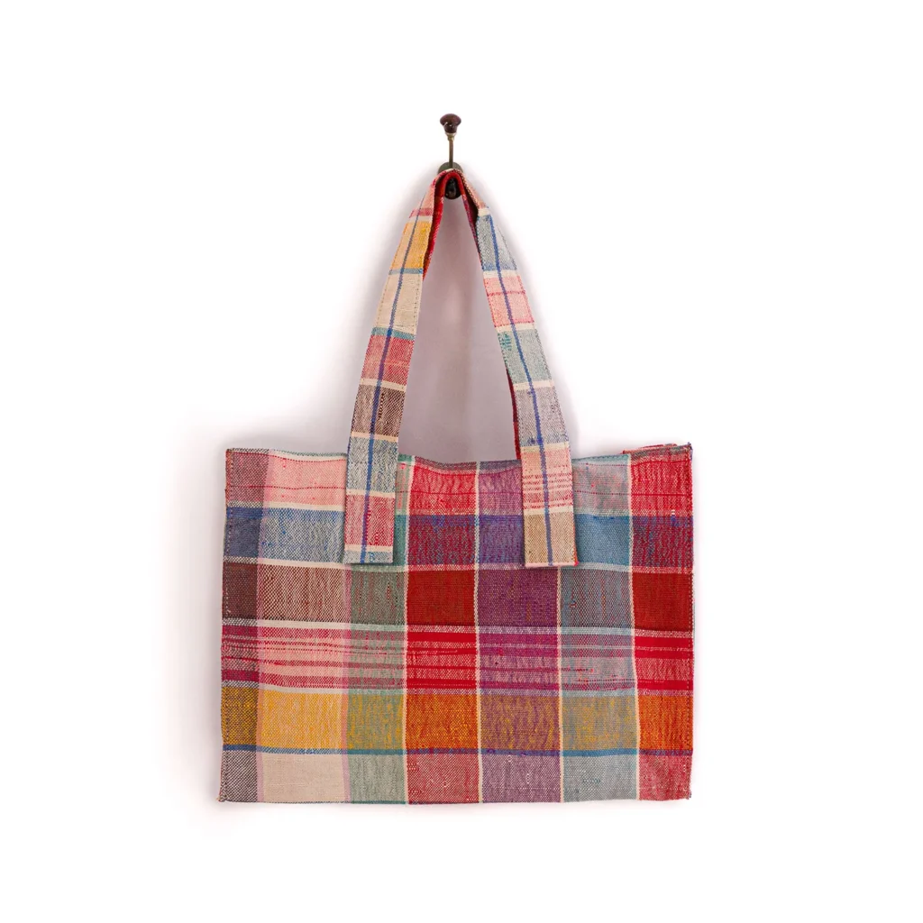 A colorful patchwork tote bag with various plaid and checkered patterns hangs against a white background.
