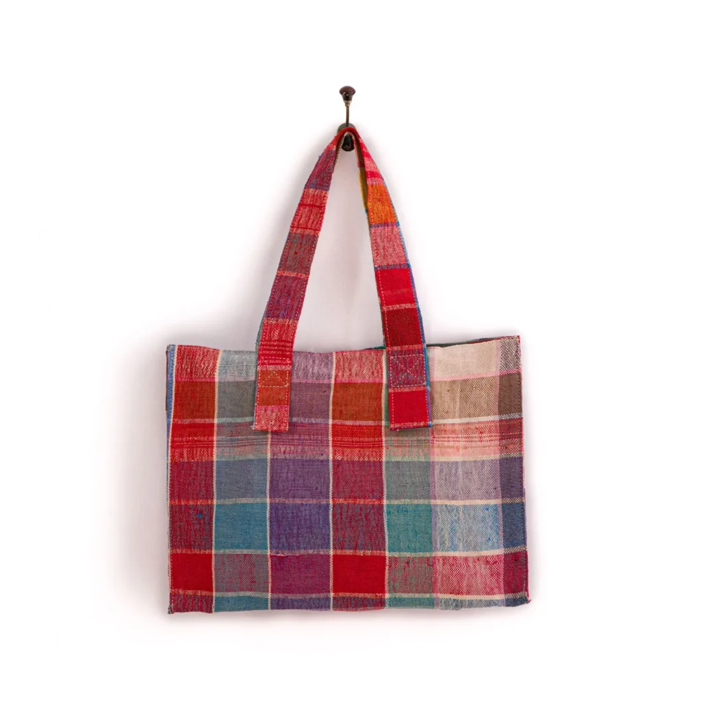 A multicolored patchwork tote bag with a plaid pattern hangs on a white wall by a hook.