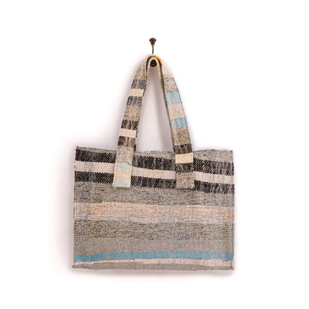 An upcycled weekend bag, this woven tote features a striking striped pattern in black, beige, blue, and orange hues, elegantly hanging on a wall hook.