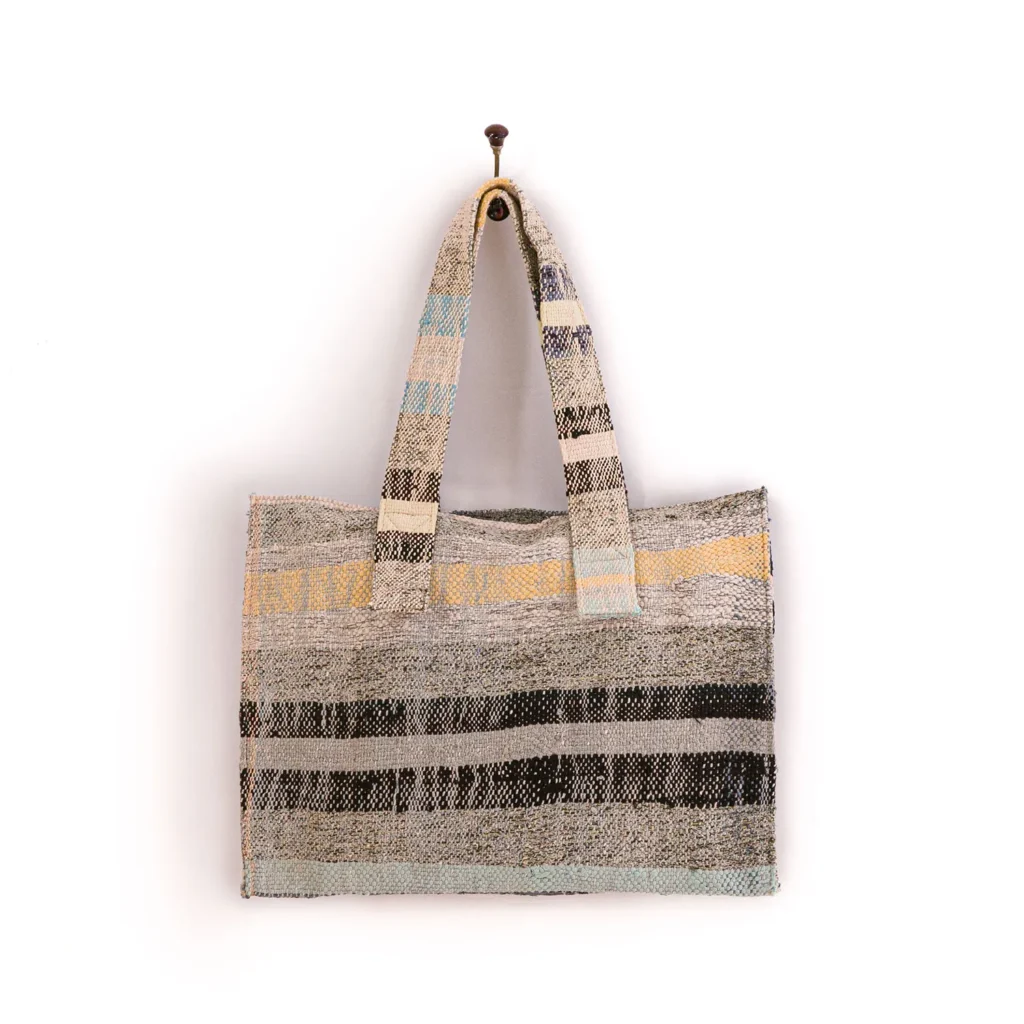 An upcycled woven tote bag with striped patterns in shades of gray, black, and muted yellow hangs elegantly on a wall hook, perfect for a relaxed weekend getaway.