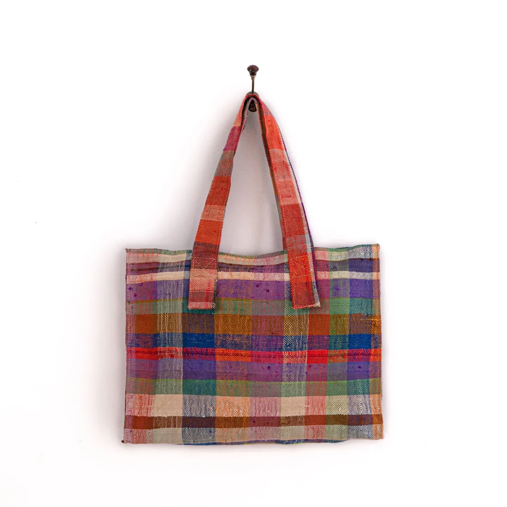 Multicolored plaid fabric tote bag with red handles, hanging on a wall hook against a white background.