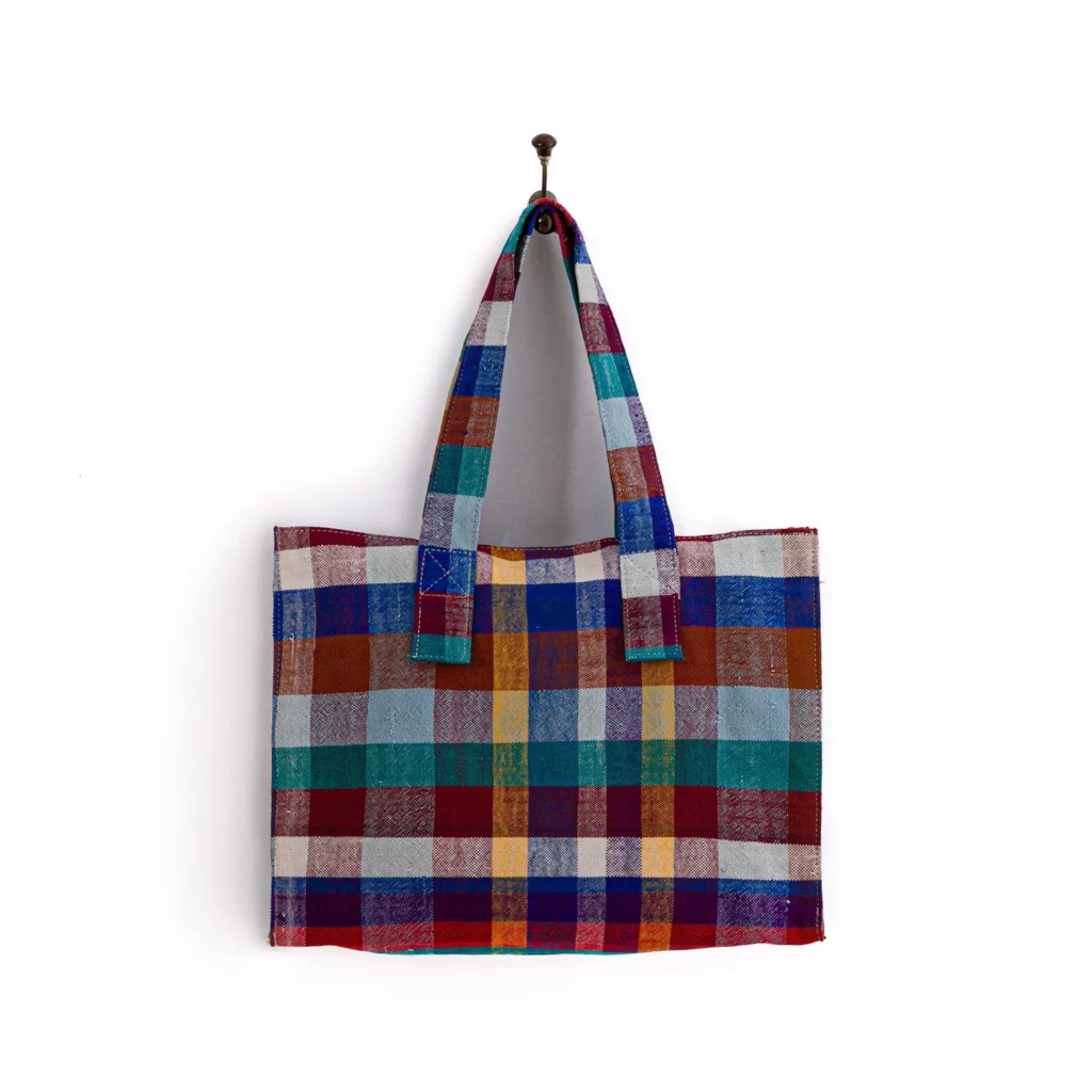 A multicolored plaid tote bag with red, blue, green, and yellow stripes hangs from a hook against a white background.