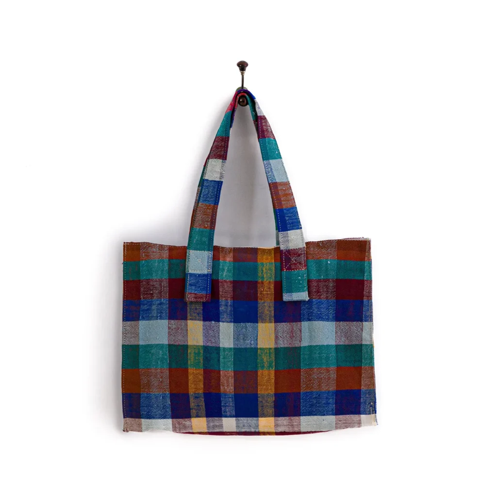 A multicolored plaid tote bag with wide handles hangs on a wall hook.