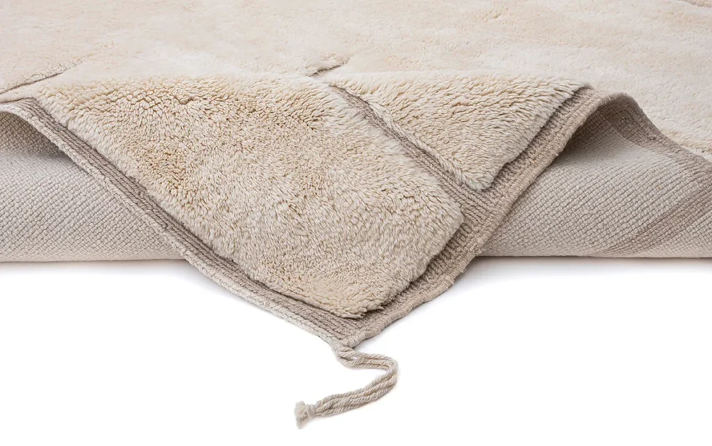 This luxurious, hand-knotted beige rug boasts a textured, fluffy top side and a smooth, woven underside. Its partially rolled design features a loose corner, adding an elegant touch to any room decor.
