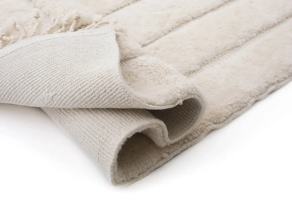 A folded beige luxury rug with a textured underside, featuring layered seams and fringed edges.